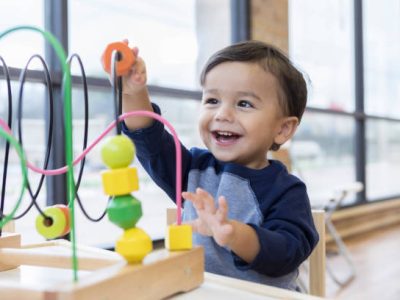 An adorable toddler boy sits at a table in a doctor's waiting room and reaches up cheerfully to play with a toy bead maze.