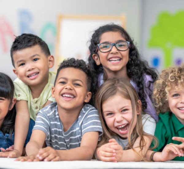 Portrait of a happy multi-ethnic group of kindergarten age students. The cute children are laying in a pile on the ground in a modern classroom. The kids are laughing and smiling.