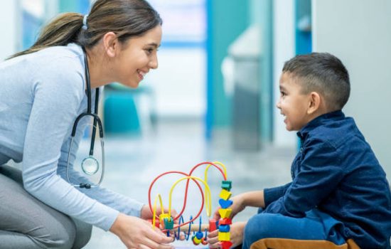 A young female, mixed race, Play Therapist, kneels down on the floor of a hospital in front of her young patient.  She is dressed semi-casually in dress pants and a button-up blouse, and has a stethoscope around her neck.  The young boy is seated cross-legged on the floor as he plays with a bead maze and talks with the therapist.