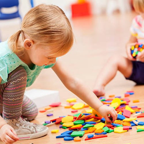 Two sweet Caucasian toddler girls playing with puzzle pieces on the floor of their preschool classroom. The puzzle pieces are colorful and the preschoolers are focused on the task at hand.